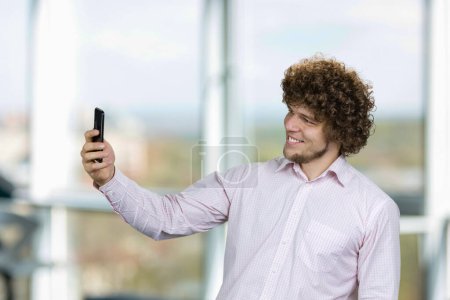 Photo for Portrait of happy young man with curly hair taking selfie with smartphone. Indoor window in the background. - Royalty Free Image