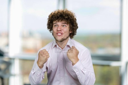 Photo for Portrait of happy excited young man with curly hair rejoicing success. Young football fan. Indoor window in the background. - Royalty Free Image