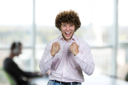 Photo for Portrait of happy excited young man with curly hair celebrates success. Indoor office interior in the background. - Royalty Free Image
