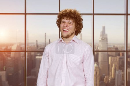Foto de Retrato de un joven feliz riendo con el pelo rizado. Ventana a cuadros con vista al paisaje urbano. - Imagen libre de derechos