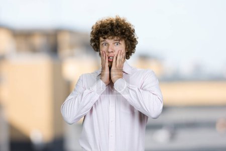 Photo for Portrait of shocked amazed young guy with curly hair touching his face. Blurred urban background. - Royalty Free Image
