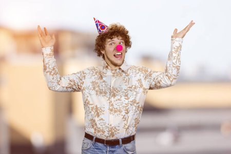 Téléchargez les photos : Un jeune homme heureux avec un nez de clown qui fait des grimaces drôles. Paysage urbain flou en arrière-plan. - en image libre de droit