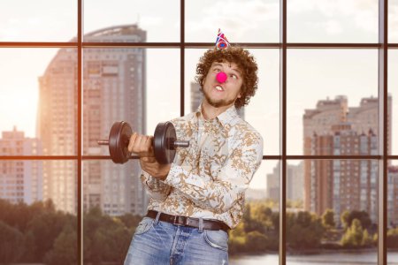 Photo for Portrait of a young clown fooling around with dumbbell indoors. Checkered window in the background. - Royalty Free Image