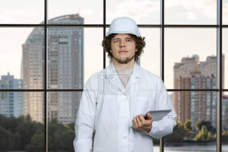 Foto de Retrato de un trabajador de la construcción joven en uniforme blanco que sostiene el dispositivo de la PC de la tableta. Ventana a cuadros con vista de paisaje urbano en el fondo. - Imagen libre de derechos