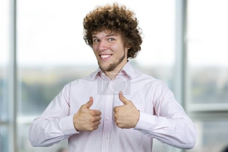 Photo for Happy smiling young man with curly hair shows both thumbs up. Indoor window in the background. - Royalty Free Image