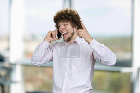 Téléchargez les photos : Portrait d'un jeune homme heureux aux cheveux bouclés parlant au téléphone se réjouissant du succès. Fenêtre intérieure en arrière-plan. - en image libre de droit