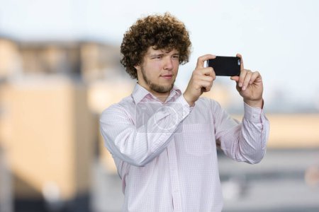 Téléchargez les photos : Portrait d'un jeune touriste aux cheveux bouclés faisant une photo sur son appareil photo smartphone. Paysage urbain flou en arrière-plan. - en image libre de droit