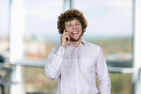 Photo for Portrait of a young man in white shirt talking on the phone and laughing. Indoor window in the background. - Royalty Free Image