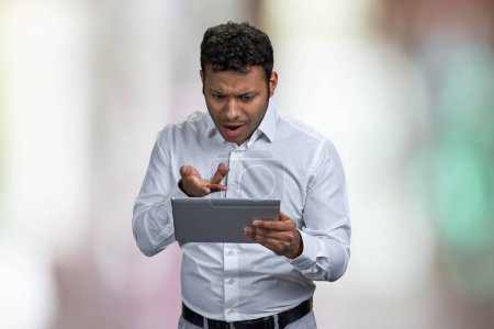 Photo for Shocked young businessman looking at digital tablet pc. Surprised man with computer tablet on abstract bokeh background. - Royalty Free Image
