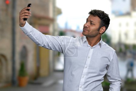Téléchargez les photos : Jeune bel homme prenant selfie avec smartphone sur fond de rue flou de la ville. Concept de personnes, technologie et mode de vie. - en image libre de droit