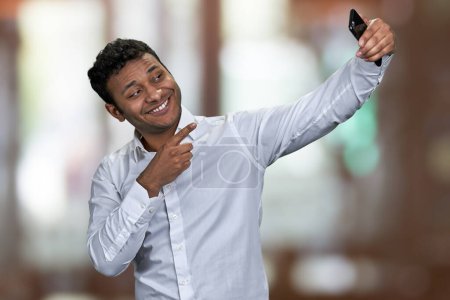 Téléchargez les photos : Joyeux jeune homme portant une chemise blanche gesticulant index à la caméra tout en prenant selfie avec son smartphone. Résumé fond bokeh. - en image libre de droit