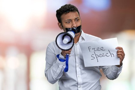 Foto de Joven empresario indio con la boca sellada sosteniendo cartel con inscripción Libertad de expresión. Fondo bokeh abstracto. - Imagen libre de derechos