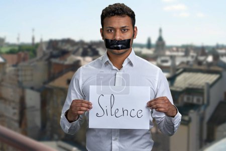 Téléchargez les photos : Homme indien à la bouche collée tenant affiche avec inscription Silence. Flou bâtiments de la ville en arrière-plan. - en image libre de droit