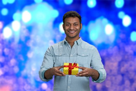 Joli jeune homme souriant qui remet un coffret cadeau à la caméra. Fond bleu bokeh lumières. Livraison du cadeau de Noël à temps.