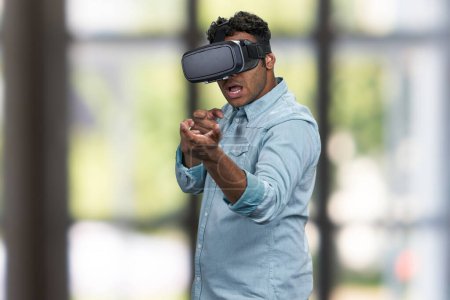Téléchargez les photos : Homme enthousiaste dans des lunettes VR simulant la prise de vue avec un pistolet virtuel. Fond flou intérieur. - en image libre de droit