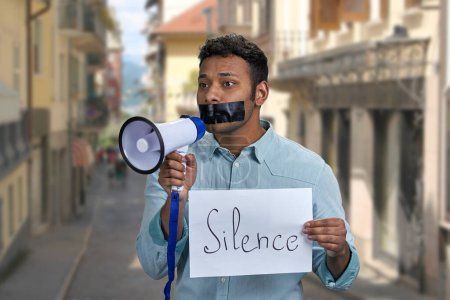 Young indian man with taped mouth unable to speak into megaphone. Censorship concept.