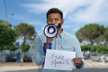 Young man with taped mouth trying to speak into megaphone standing outdoors. Free speech concept.
