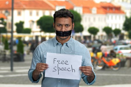Photo for Young indian man with taped mouth holding paper card with handwritten inscription Free speech. Blur city street in the background. - Royalty Free Image