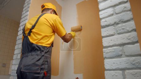 Foto de El trabajador pinta el rodillo de pared. El pintor de la casa pinta la pared en marrón. - Imagen libre de derechos