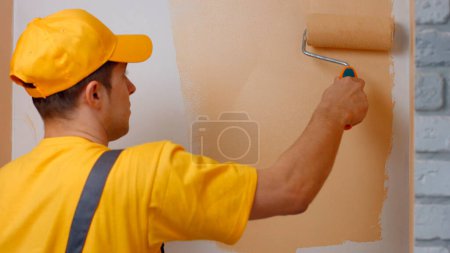 Téléchargez les photos : Builder peint un mur blanc de couleur beige avec un rouleau. Peinture murale professionnelle. - en image libre de droit