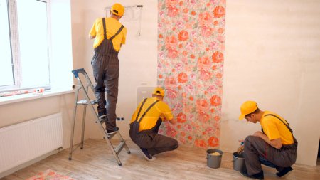 Foto de Brigada de constructores cuelgan papel pintado en el apartamento. Maestros hacen reparaciones. - Imagen libre de derechos