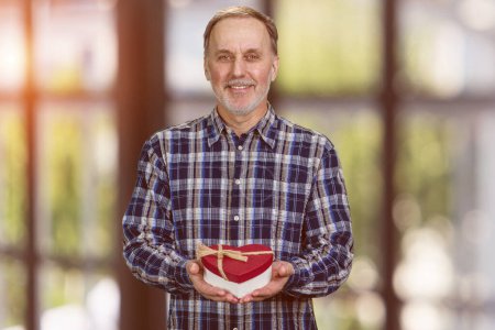 Photo for Happy smiling handsome senior man is giving a heart shape gift box. Valentines day or anniversary. Blurred window background. - Royalty Free Image