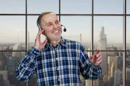 Photo for Portrait of smiling mature male call center customer support worker. Checkered window backgroud with cityscape view. - Royalty Free Image