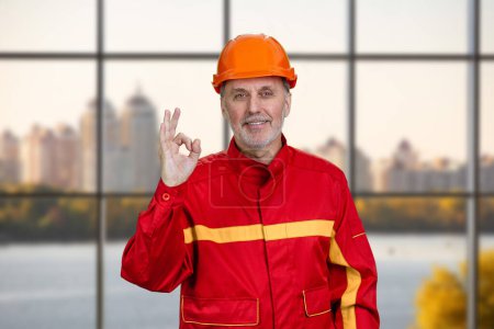 Joyeux travailleur de la construction souriant en uniforme rouge montre signe de geste correct. Fond de fenêtre à carreaux.