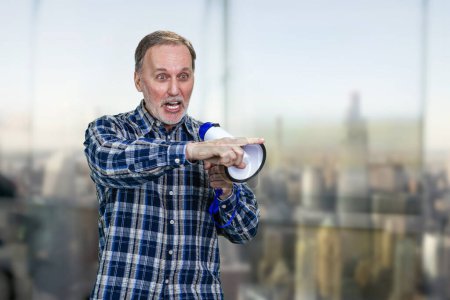 Confident elderly mature man giving a speech in megaphone and pointing at something. Boss giving a command. Blurred indoors background.