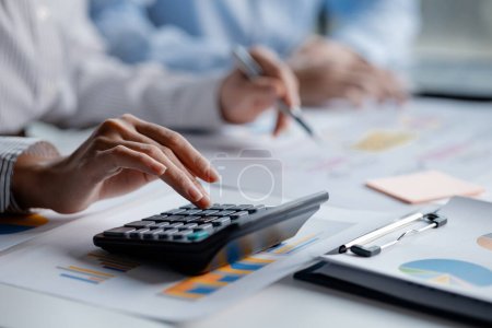 Photo for Businessman is using a calculator to calculate company financial figures from earnings papers, a businessman sitting in his office where the company financial chart is placed. - Royalty Free Image