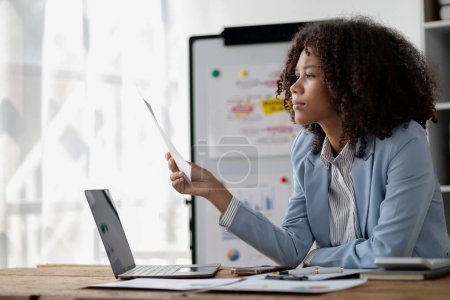 Photo for American woman sitting working in a startup company, american businesswoman working in the office, she is looking at financial documents and sales data to summarize before meeting with executives. - Royalty Free Image