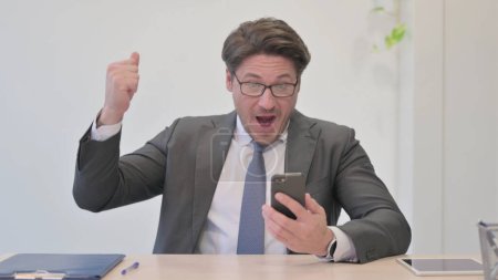 Téléchargez les photos : Homme d'affaires du Moyen Age célébrant le succès en ligne sur Smartphone au bureau - en image libre de droit