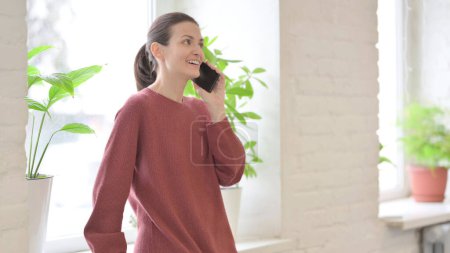 Téléchargez les photos : Jeune femme parlant au téléphone tout en se tenant près de la fenêtre - en image libre de droit