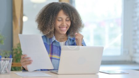 Téléchargez les photos : Jeune femme africaine célébrant le succès avec ordinateur portable et documents, Contrat - en image libre de droit