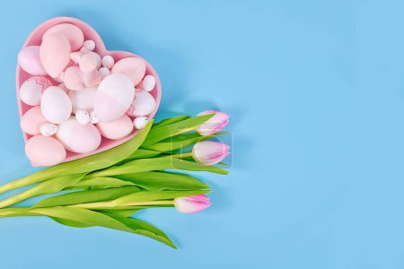 Easter decoration with painted eggs in heart shaped plate and pink tulip flowers on blue background with copy space