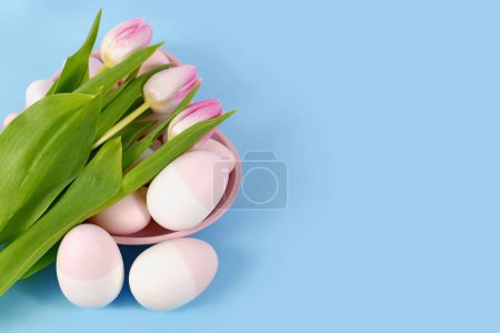 Easter decoration with painted eggs in heart shaped plate and pink tulip flowers on blue background 