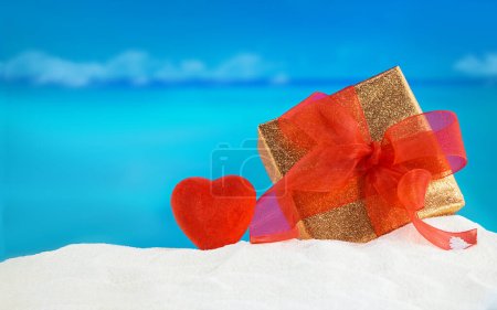 Foto de Caja de regalo de oro con lazo rojo y corazón en la playa de arena, con el mar detrás. Vacaciones, relajación, viajes a países calientes. San Valentín, Día de la Madre y de la Mujer, Navidad, Año Nuevo, cumpleaños, felicitaciones, vacaciones. Copiar espacio - Imagen libre de derechos