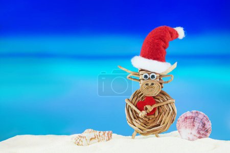 Foto de Vaca tejida de juguete con sombrero de Santa con corazón y conchas en la playa de arena con el mar. Navidad, Año Nuevo, San Valentín. Países calientes. Copiar espacio - Imagen libre de derechos