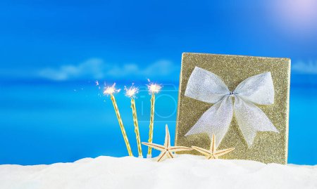 Photo for Golden shiny box with silver bow, candles with flames and starfish on sand of beach. Christmas, New Year, birthday, International Women's and Mother's Day, holiday. In hot countries. Copy space - Royalty Free Image