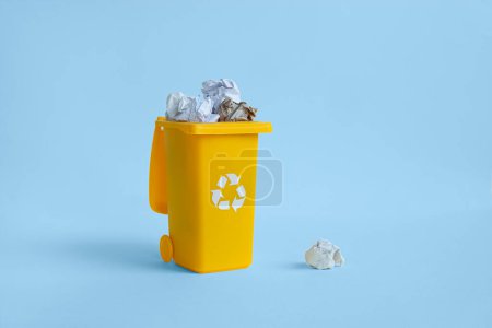Geöffneter gelber Behälter für die Papiermüllabfuhr mit zerknülltem Papier im Inneren, isoliert auf blauem Hintergrund mit Kopierraum, Abfallrecycling-Konzept