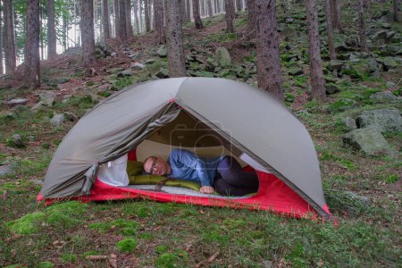 Foto de Caminante con rastas acostado en la almohadilla y saco de dormir en verde tienda de campaña de 2 paredes por la mañana en el bosque en las montañas Beskid - Imagen libre de derechos