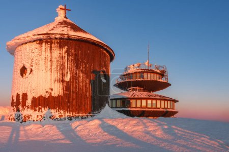 Winter, sunrise time, wooden Roman catholic chapel and disc shaped meteorological observatory in snezka, mountain on the border between Czech Republic and Poland.