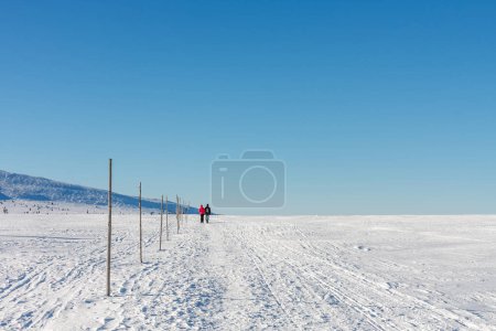 Téléchargez les photos : Deux touristes marchant dans les montagnes krkonose, chemin de la maison silésienne à Meadow Hut, matin d'hiver. Le long de la route se trouvent de longs bars en bois, des marques touristiques pour la saison d'hiver. - en image libre de droit