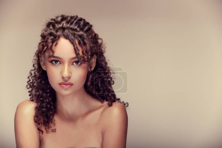 Photo for "Image of a beautiful woman, with a shot capturing her mid-body, her curly hair, and a penetrating gaze, all framed against a beige background - Royalty Free Image
