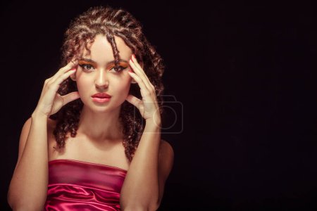 Photo for Medium shot portrait of a woman in a red dress against a black backdrop, with wavy hair, red makeup, and hands delicately placed beside her face - Royalty Free Image