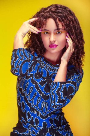 Photo for Young woman with an intense gaze and hands on her face, wearing a blue dress, and yellow background - Royalty Free Image
