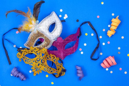 Photo for Three masks together, one golden, one violet, and one white, with feathers, isolated on a blue background - Royalty Free Image