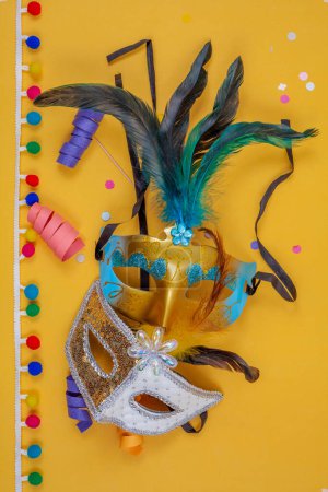 Photo for Two carnival masks together adorned with feathers, confetti, and colorful streamers on a vertical yellow background - Royalty Free Image