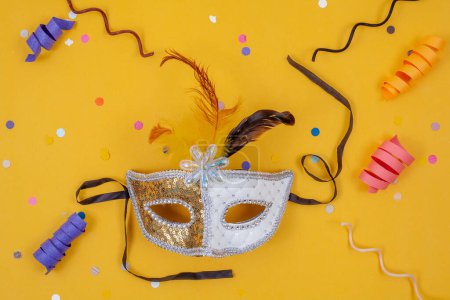 Photo for A white and gold mask stands alone against a vibrant yellow backdrop, adorned with a festive display of colorful confetti and streamers - Royalty Free Image