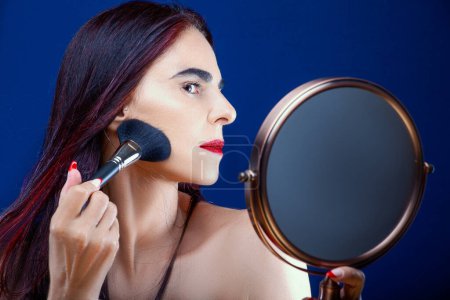 Photo for Woman applying makeup reflected in a mirror against a blue background - Royalty Free Image